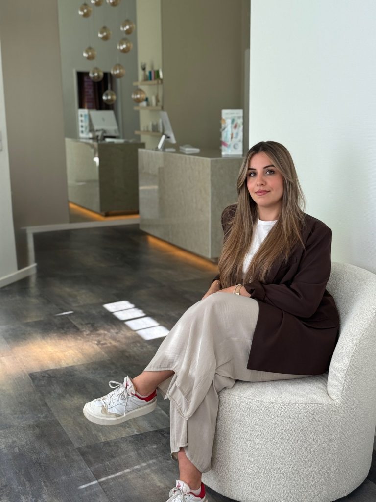 Eine Person mit langen, braunen Haaren sitzt auf einem hellen Sessel in einem modernen Raum.