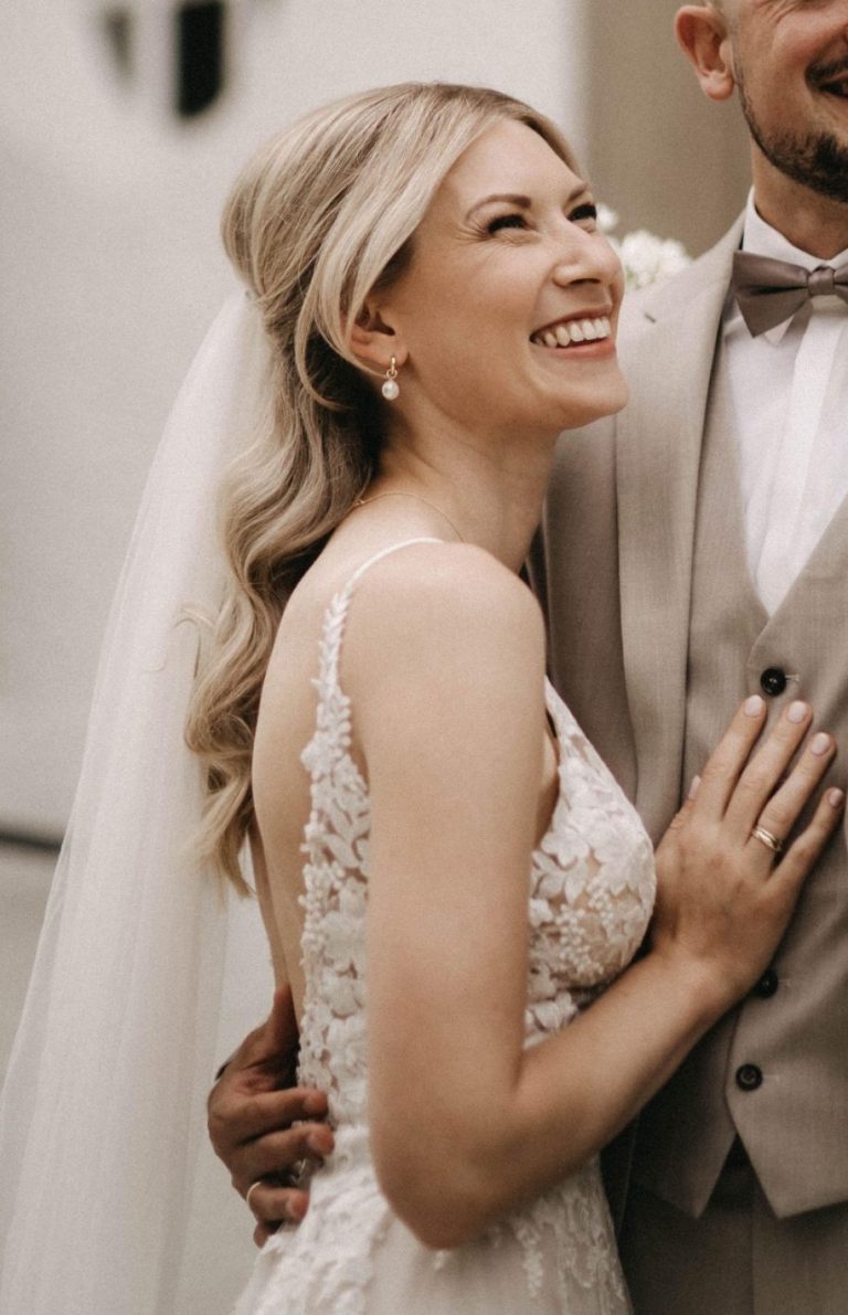 Braut in einem eleganten Kleid mit Blumenarrangement, lächelnd und geschminkt von ALMA Kosmetik & Wellness.