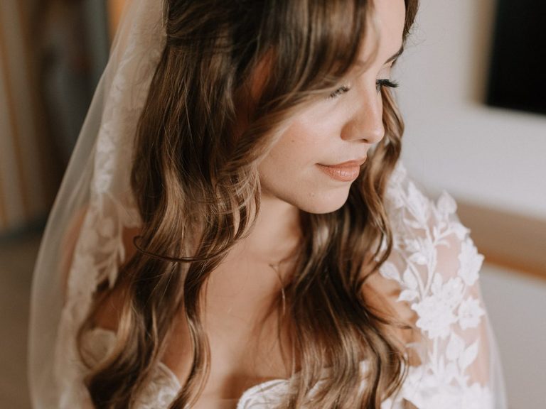 Braut mit langen, lockigen Haaren und zartem Schleier, sieht nachdenklich aus. Geschminkt von ALMA Kosmetik & Wellness